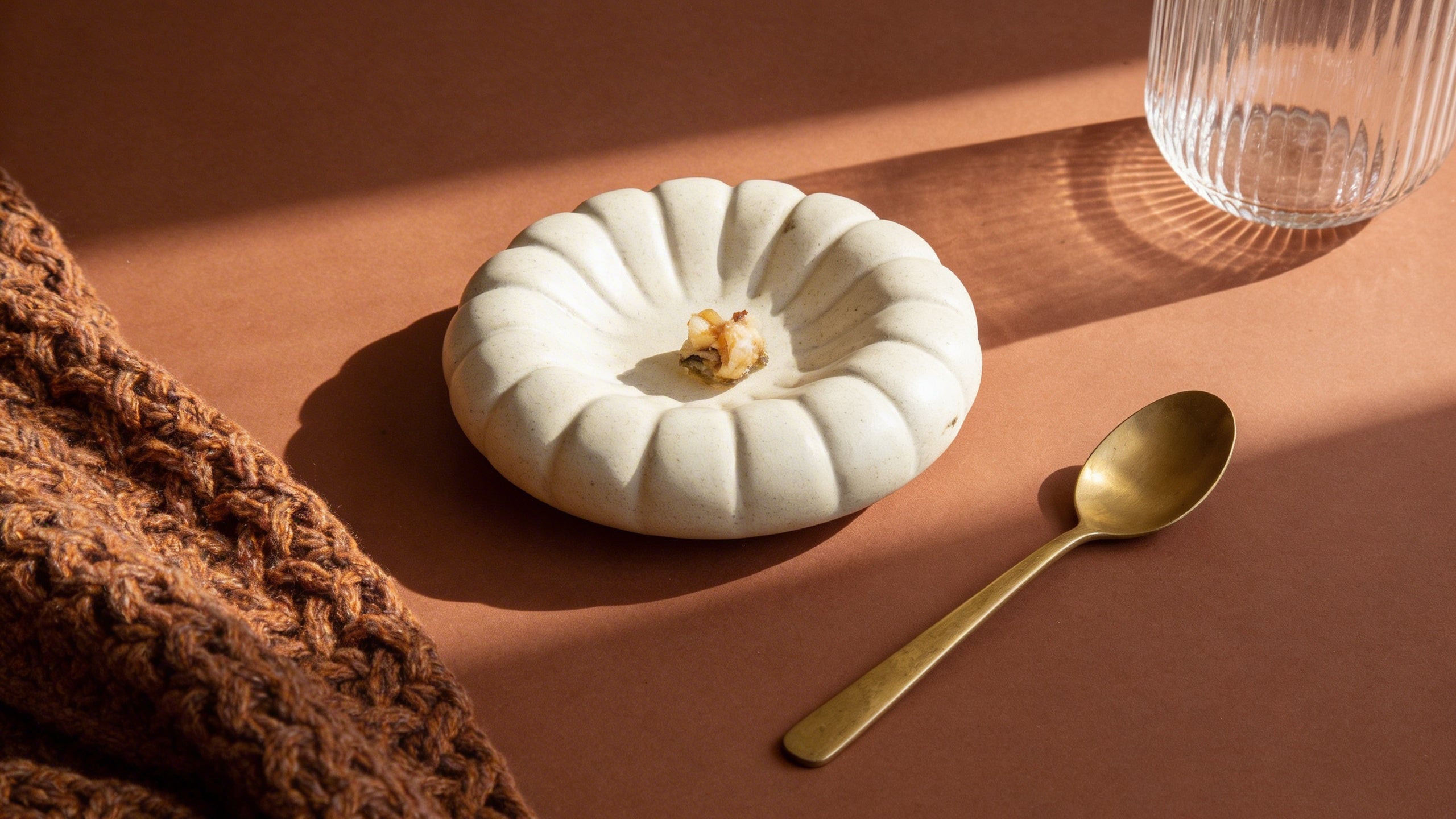 A white ceramic pumpkin-shaped dish with a small food piece in the center, styled on a warm brown surface with a gold spoon, a glass, and a chunky brown knit textile in soft sunlight.
